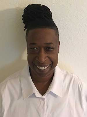 A Black person wears a white shirt while standing in front of a white wall. Their hair is braided and pulled back into a bun at the crown of their head. They're smiling.