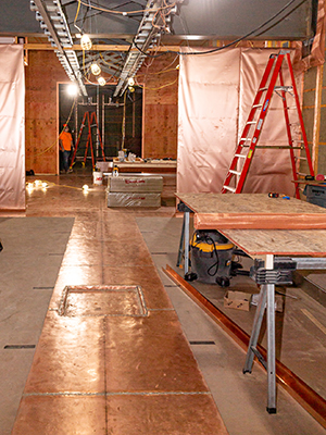 A room shows large sheets of copper covering walls and floors. There is construction present in the photo. A red ladder sits off to the right. A construction bench is also in the foreground.