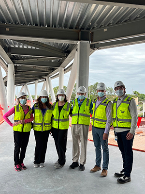 6 people wear hard hats, neon green reflector vests and masks as they stand on a semi-completed pedestrian bridge. Cloudy skies and a construction zone is somewhat visible in the background.