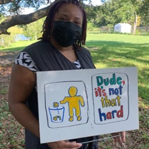 A woman wearing a black mask holds a white sign. The sign shows a figure painted in yellow placing trash into a bin. Beside the figure, it says "Dude, it's not that hard."