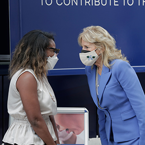 Jackie Smith (left) wears a white blouse and white mask while speaking with Dr. Jill Biden (right) wearing a blue suit and white mask. They're standing in front of a blue bus with signage about cancer screenings in the background.