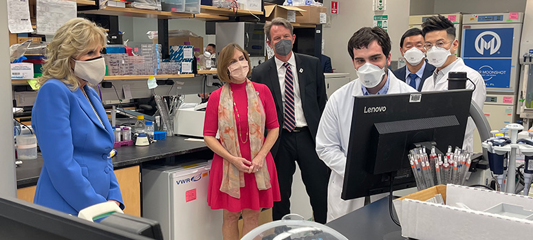Dr. Biden (left) wears a blue suit and white mask while visiting a research lab at Moffitt Cancer Center. She is joined by U.S. Representative Kathy Castor, NCI Director Ned Sharpless, cancer biology student Chase Burton, Dr. Patrick Hwu and Dr. Eric Lau. She group is looking at a computer. There is lab and research equipment in the background.