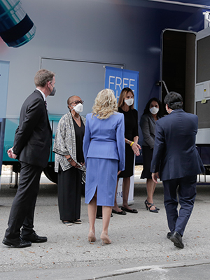 Dr. Biden has her back to the camera and walks toward a bus with screening flags in front of it. Accompanying her are Dr. Ned Sharpless and Dr. Patrick Hwu. Waiting by the bus to greet her are the mother of Jackie Smith, Dr. Jane Messina and Dr. Susan Vadaparampil.